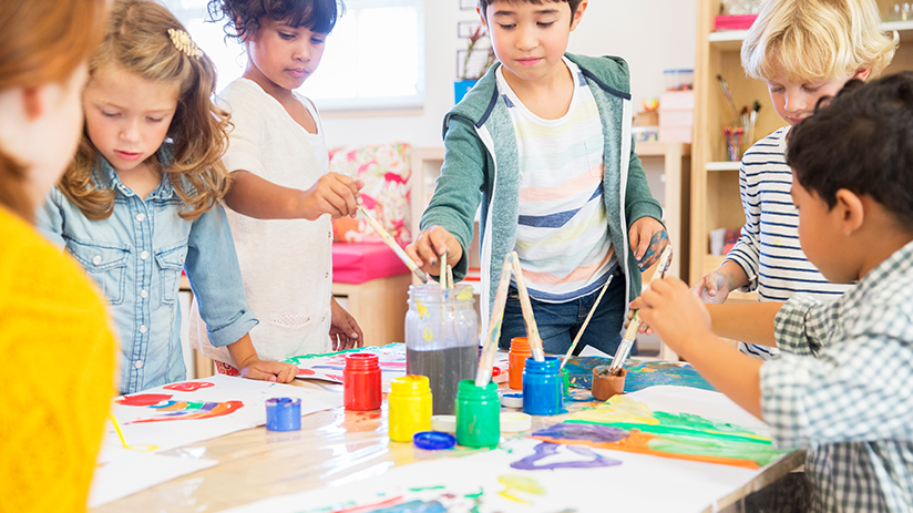 Eine Gruppe Kindergartenkinder steht um einen Tisch und malt mit Wasserfarben.