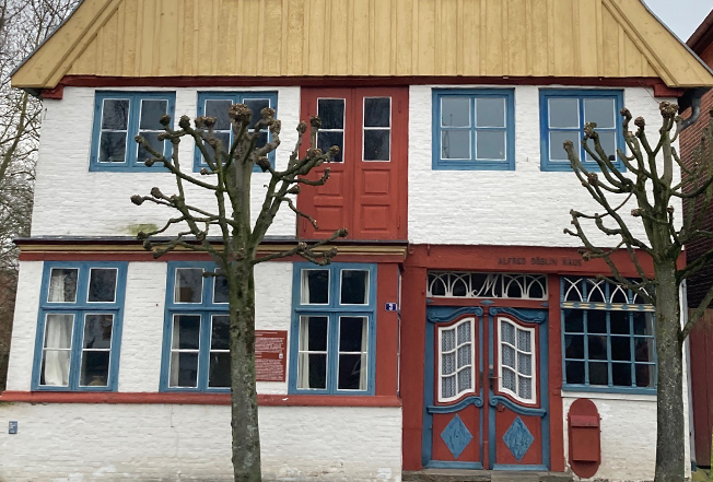 Frontalansicht des Hauses mit weißer Hauswand, blauen Fensterrahmen und roten Türen, der Dachgiebel ist aus hellem Holz
