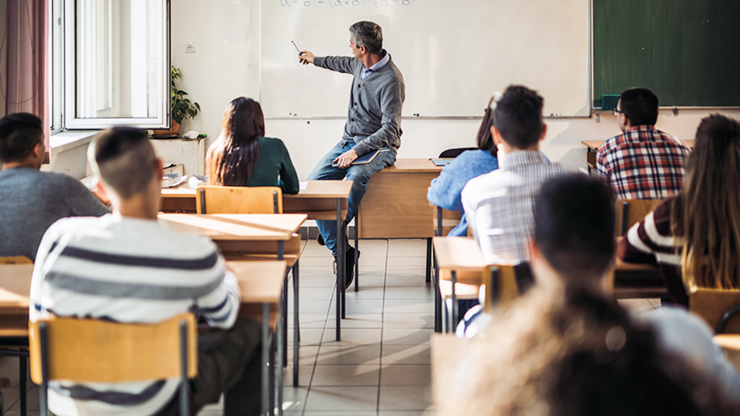 Ein Lehrer steht vor einer Klasse Jugendlicher und zeigt auf die Tafel hinter sich. 