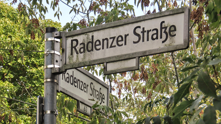 Straßennamensschild mit Radenzer Straße darauf geschrieben. Rund ums Schild stehen Bäume.